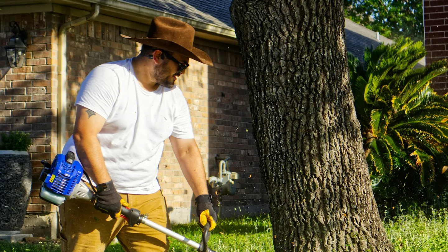 Hombre cortando hierba junto a un árbol con una desbrozadora