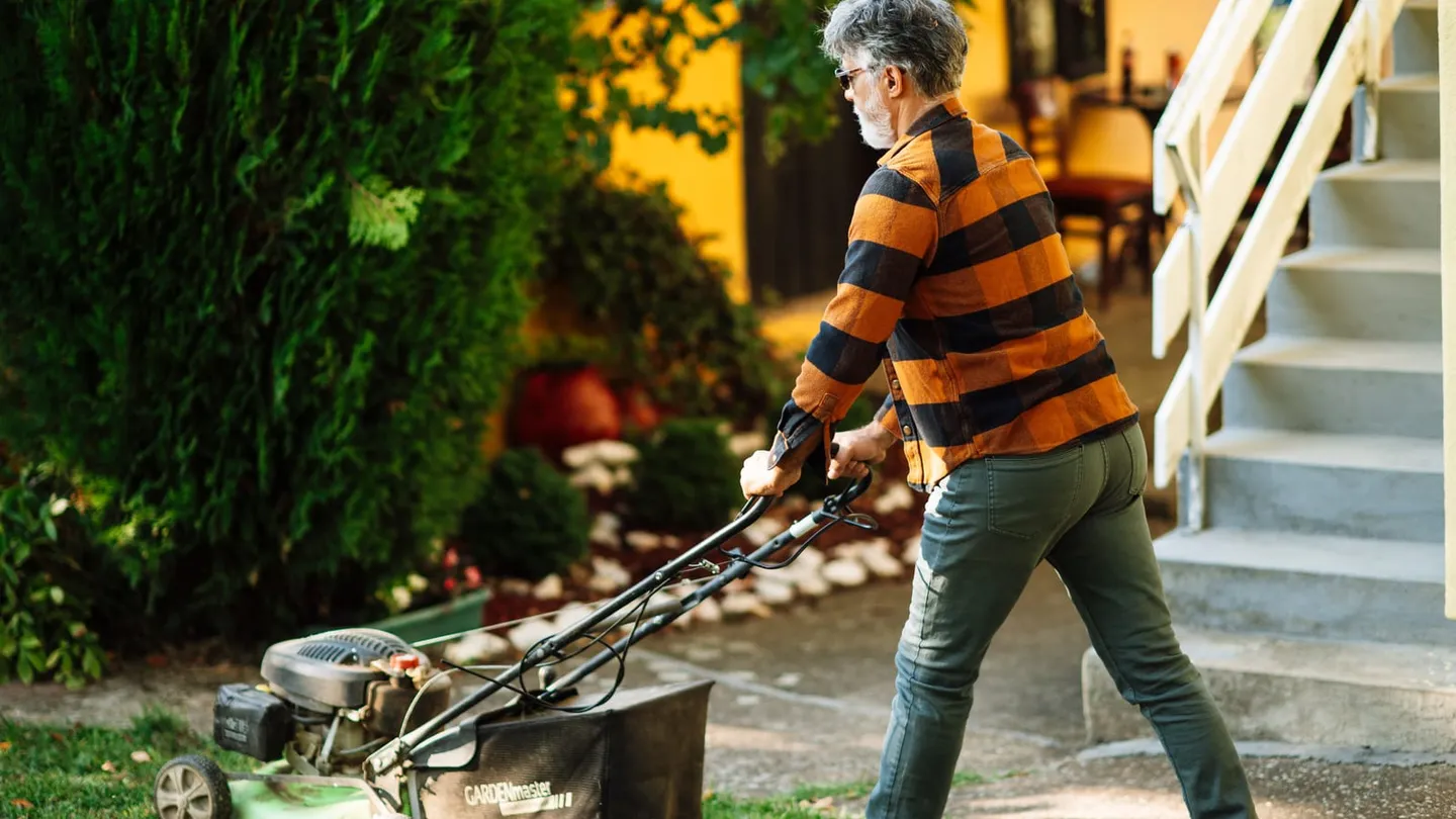 Hombre con tenis y pantalón largo empujando una podadora de gasolina en el jardín